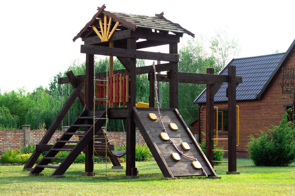 Paisagem. Parque infantil de madeira com baloiços, corrediça e escadas no fundo de uma cerca de tijolo no pátio da casa particular. - Foto, Imagem