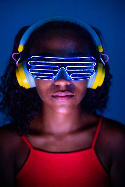 Junge Frau mit Kopfhörern und futuristischer LED-Brille auf blauem Hintergrund - Vereinzelte schwarze Frau mit 3D-Smart-Brille und Kopfhörer - Virtuelle Realität, Zukunft, Technologiekonzept  - Foto, Bild