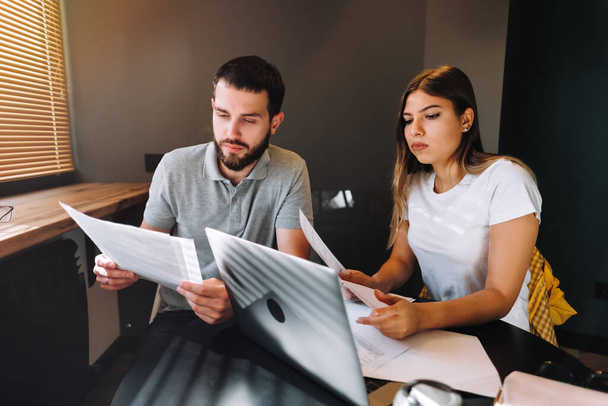 A young couple manages finances by looking through their bank accounts in the living room at desk using laptop. Woman and man look at documents together. Planning budget expenses. High quality photo - Foto, Bild
