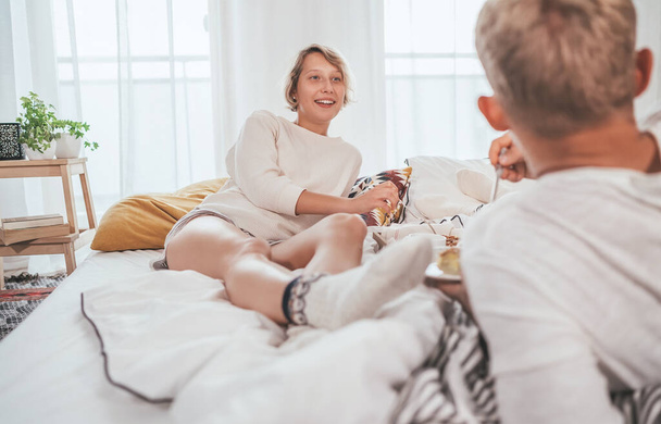 Laughing blonde hair young adults couple in pajamas lazy relaxing lying in a cozy bed in the bedroom and having a peacefully carefree chatting. Couples relations concept image. - Фото, изображение