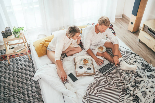 Top view of the couple in pajamas in cozy bed browsing internet together using a laptop and having a morning coffee with apple pie dessert. - Φωτογραφία, εικόνα