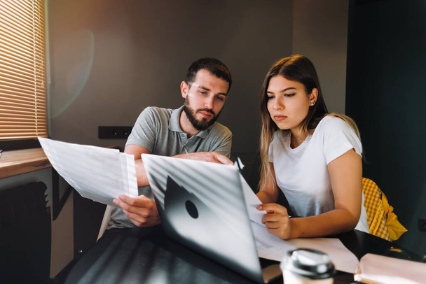 A young couple manages finances by looking through their bank accounts in the living room at desk using laptop. Woman and man look at documents together. Planning budget expenses. High quality photo - Foto, Bild
