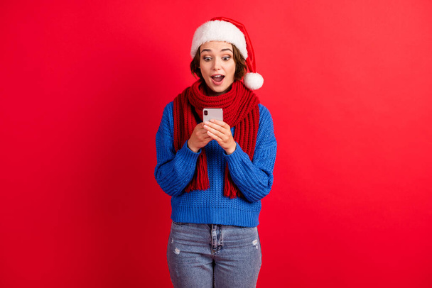 Shocked amazed girl in santa claus cap use smartphone get christmas congratulation famous social media blogger wear blue knitted sweater denim jeans isolated bright shine color background - Foto, afbeelding