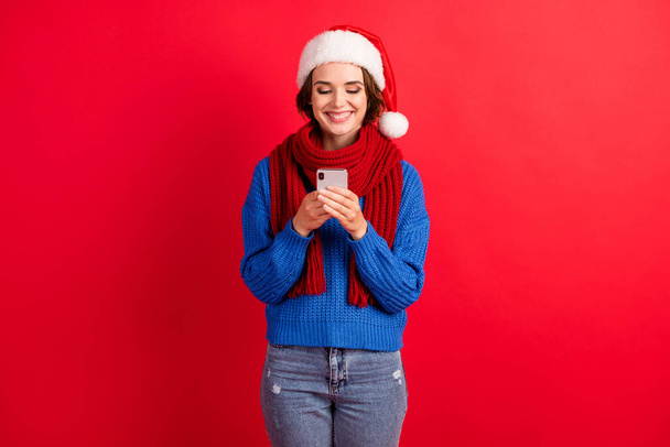 Photo of positive girl in santa claus cap use smartphone typing christmas x-mas newyear congratulations wear blue knitted sweater denim jeans isolated over bright shine color background - Фото, изображение