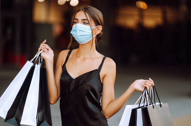 Beautiful fashionable woman  in protective  medical mask with shopping bags walking near mall. Shopping during the coronavirus Covid-19 pandemic. Purchases, black friday, discounts, sale concept.  - 写真・画像