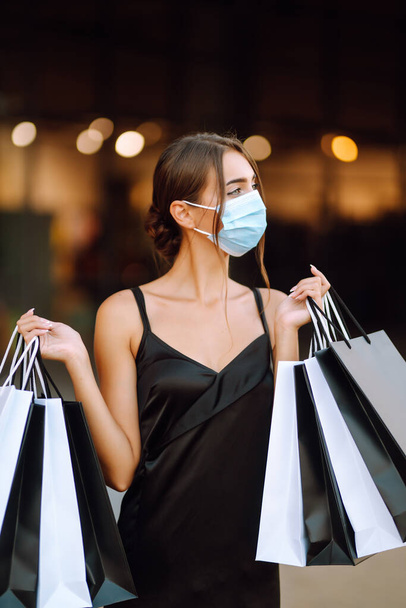 Beautiful fashionable woman  in protective  medical mask with shopping bags walking near mall. Shopping during the coronavirus Covid-19 pandemic. Purchases, black friday, discounts, sale concept.  - Фото, зображення