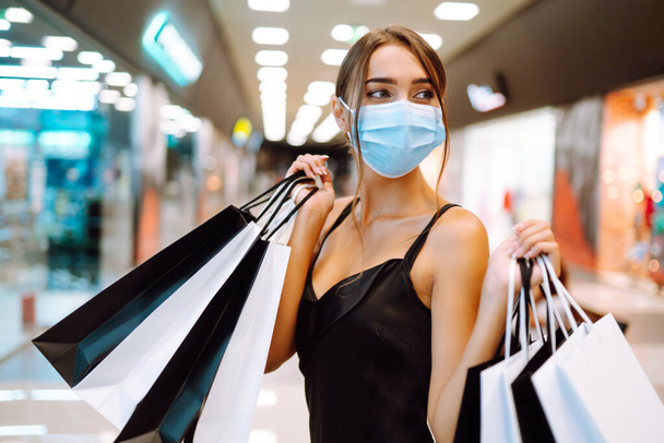 Fashionable woman in protective  medical mask with shopping bags walking in the mall. Shopping during the coronavirus Covid-19 pandemic. Purchases, black friday, discounts, sale concept.  - Fotoğraf, Görsel