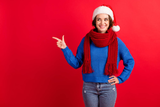 Photo positive girl in santa claus headwear point finger copyspace demonstrate christmas season tradition discount wear blue pullover denim isolated red bright shine color background - Fotografie, Obrázek