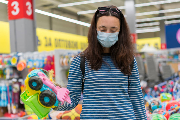 La mujer con la máscara protectora en el supermercado escoge los juguetes infantiles. Mujer con máscara médica en el supermercado. - Foto, Imagen