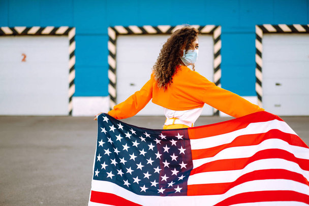 Koruyucu maske takan ve sokak şehrinde ABD bayrağı taşıyan genç bir kadının portresi. Özgürlük ve anıt konsepti. Covid 2019. - Fotoğraf, Görsel
