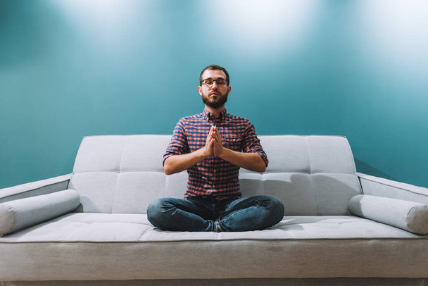 Young caucasian man with eyes closed sitting on couch and meditating at home. High quality photo - Photo, image