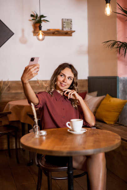 Hymyilevä houkutteleva nuori valkoihoinen nainen tekee selfien älypuhelimellaan ja pitämällä kupin kahvia. Nykyaikaisessa hipsterikahvilassa on päiväsaika. Vaikutus bloggaaja käsite. - Valokuva, kuva
