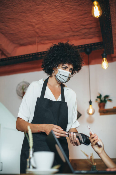 Waitress wearing a protective face mask, and showing the card terminal to a customer during Coronavirus pandemic. Concept online shopping, wallet app, wireless nfc technology. - Photo, Image