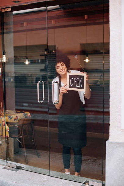 Portrait of beautiful business woman, with afro hairstyle, putting an open sign on the glass door. Happy female owner attending new customers. Small business concept, restaurants, birth of new store. - Photo, image