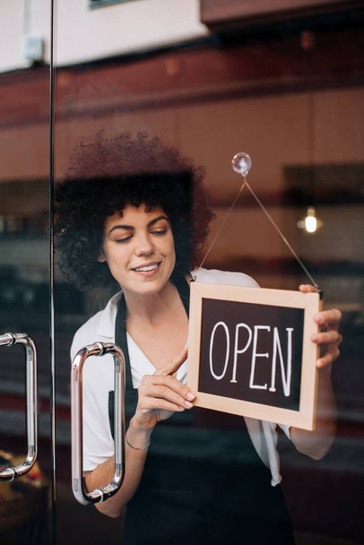 Portrait of beautiful business woman, with afro hairstyle, putting an open sign on the glass door. Happy female owner attending new customers. Small business concept, restaurants, birth of new store. - Foto, Bild
