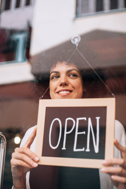 Portrait of smiling business woman, with afro hairstyle, putting an open sign on the glass door. Happy female owner attending new customers. Small business concept, restaurants, birth of new store. - Фото, изображение