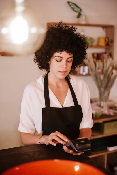 Portrait of a caucasian female barista working with the card terminal. Woman with apron standing behind the bar counter. Concept small business owner, young entrepreneur. - Foto, imagen