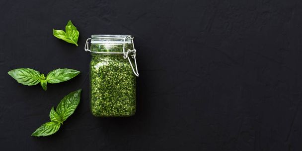 Homemade pesto sauce and green basil on dark black background. Italian green pesto sauce in glass jar. Vegetarian healthy food. Top view long web banner with copy space for text - Fotó, kép