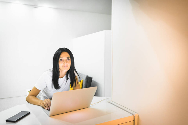 Nuori kaunis valkoihoinen nainen istuu työpöydällä kannettavalla tietokoneella ja näyttää suoraan valkoisessa valoisassa huoneessa. Työ kotikonseptista taustakuva. - Valokuva, kuva
