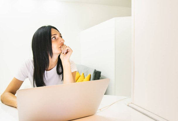 Jeune femme brunnete assis derrière un ordinateur portable et regarde vers le haut d'être attentionné. Concept d'études et de travail à domicile. - Photo, image