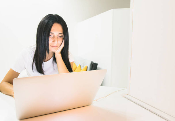 Primer plano de mujer joven caucásica en camisa blanca con el pelo negro sentado relajado y mirando a la pantalla de la computadora en la habitación del hogar brillante. - Foto, imagen
