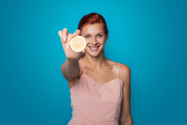 La donna dai capelli rossi sta mostrando alla telecamera un limone affettato che promuove uno stile di vita sano su una parete blu dello studio - Foto, immagini