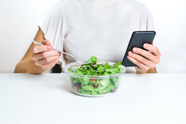 Eine bis zur Unkenntlichkeit kaukasische junge Frau bedient sich eines Smartphones, während sie einen Salat aus einer Lunchbox isst. Konzept der Sucht nach neuen Technologien - Foto, Bild