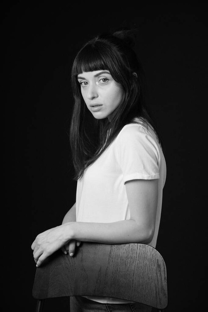 Studio portrait of a pretty brunette woman in a white blank t-shirt, standing behind the chair, against a plain black background, looking at camera - Foto, imagen