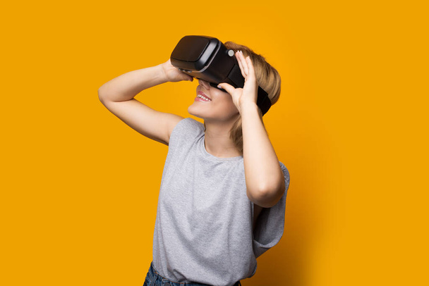 Blonde caucasian lady looking up at something using a virtual and augmented reality headset posing on a yellow wall - Photo, Image