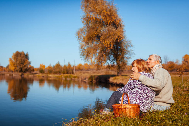 Starsza rodzina urządza piknik nad jesiennym jeziorem. Szczęśliwy mężczyzna i kobieta cieszący się naturą i przytulaniem. Romantyczna data - Zdjęcie, obraz
