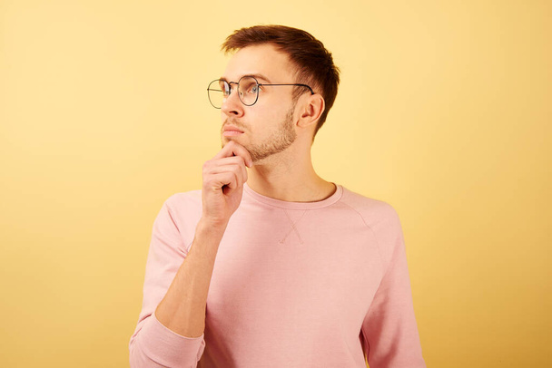 Portrait of a guy wearing glasses with a pensive face, holding a chick and thinking, dressed in pink casual isolated on yellow background - Foto, Imagen
