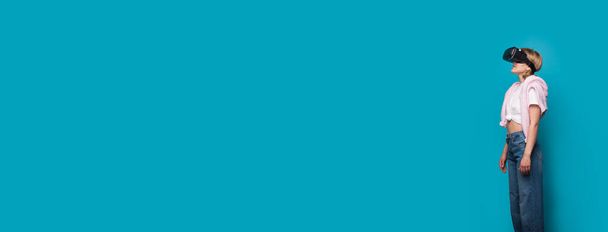 Mulher caucasiana loira com um fone de ouvido realidade virtual olhando para o estúdio azul livre espaço em branco anunciando algo - Foto, Imagem
