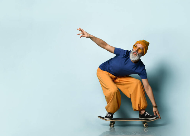 Bearded elderly man in t-shirt, sunglasses, orange pants, hat, gumshoes. Riding black skateboard, posing on blue background - Foto, Imagen