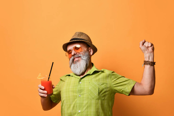 Gray-bearded man in hat, green shirt, sunglasses. Smiling, stretching himself, holding glass of juice, posing on orange background - Photo, image