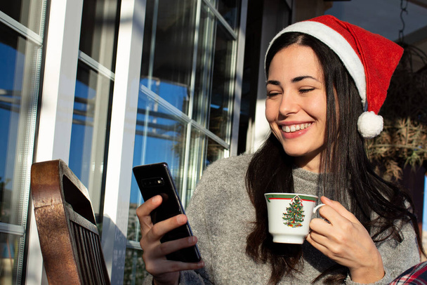 Μια όμορφη νεαρή γυναίκα χαμογελά κρατώντας ένα φλιτζάνι καφέ χρησιμοποιώντας το κινητό της, ενώ φοράει ένα καπέλο Santa Claus - Φωτογραφία, εικόνα
