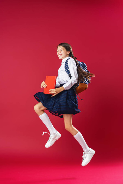 mischievous schoolgirl, in uniform with backpack and books jumps on red background. Dynamic images. go back to conceptual school. holidays begin. Back to school. girl is ready to study - Foto, immagini