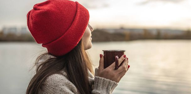 Punahattuinen nainen juo kahvia järvellä auringonlaskun aikaan. Syksyn ulkoilu. Matkustusvapauden käsite - Valokuva, kuva