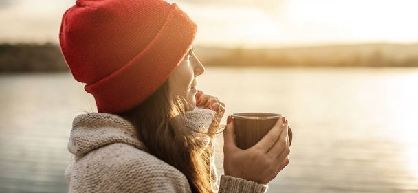 赤い帽子の女性は日没時に湖でコーヒーを飲んでいる。秋の屋外レクリエーション。旅の自由の概念 - 写真・画像