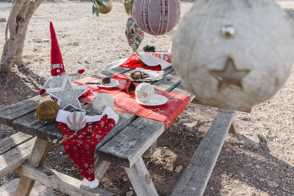 Piknik asztal karácsonyi dekorációval a mezőn, karácsonyi torta maradványaival és egy fa ágain lógó dekorációkkal. - Fotó, kép