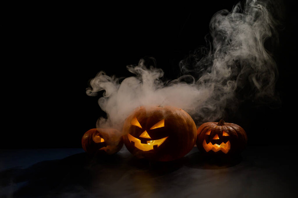 Three creepy halloween steaming pumpkins with a carved luminous smirk on a black background. A handmade jack-o-lantern head with a candle inside in the dark among the fog. Trick or treat. Postcard. - Fotografie, Obrázek