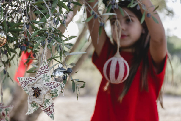 Ein unkonzentriertes Mädchen hängt Weihnachtsschmuck an den Zweigen eines Olivenbaums auf dem Feld und trägt ein Stirnband - Foto, Bild