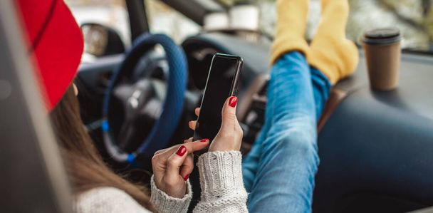 Женщина в машине в теплых носках держит мобильный телефон гаджет и чашку кофе. Уютная осенняя поездка на выходные. Концепция свободы передвижения - Фото, изображение