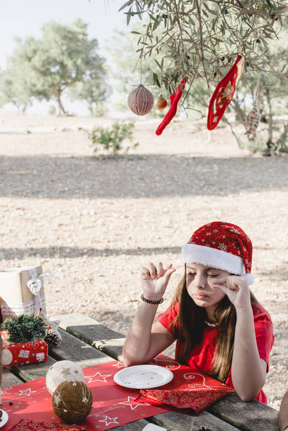 A girl celebrating a Christmas picnic in Santa hats, eating at a table under a tree with hanging Christmas decorations - Photo, Image