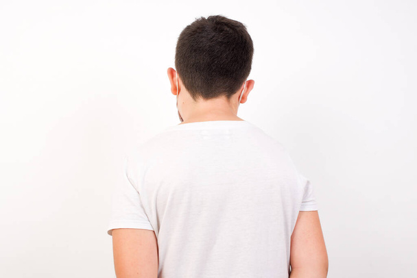 Η πίσω όψη ενός όμορφου αγοριού που φοράει καθημερινά ρούχα και στέκεται στον λευκό τοίχο. Στούντιο σουτ.  - Φωτογραφία, εικόνα