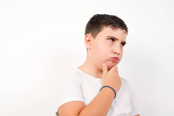 Isolated portrait of stylish young caucasian boy with hand under chin and looking sideways with doubtful and skeptical expression, suspect and doubt. Standing indoors over white background. - Фото, зображення