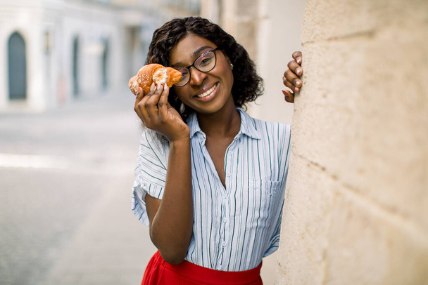 Close up urban lifestyle portrait of pretty cute African American lady, smiling to camera, funny hiding her eye with fresh croissant, standing outdoor on the background of old city buildings - Foto, Imagen