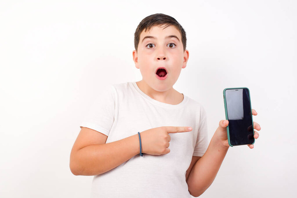 Excited boy standing against white background pointing with finger at digital device. Studio shot of shocked guy holding smartphone with blank screen. Advertisement concept.  - Foto, imagen