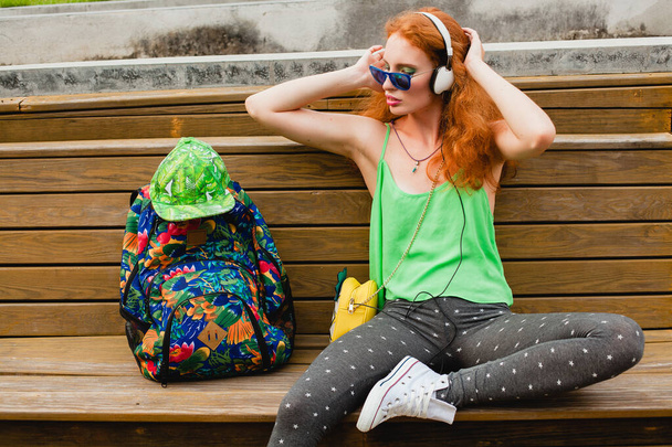 mujer joven y elegante jengibre hipster, sentado en el banco, escuchando música, auriculares, gorra verde, leggings, gafas de sol, zapatillas de deporte, divertirse, ropa de moda de moda, mochila, turista, relajarse - Foto, imagen