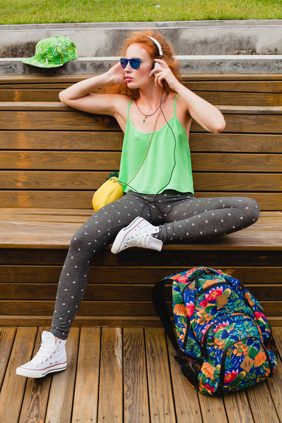 young stylish hipster ginger woman, sitting on bench, listening music, headphones, green cap, leggings, sunglasses, sneakers, having fun, trendy fashionable outfit, backpack, tourist, relax - Foto, imagen
