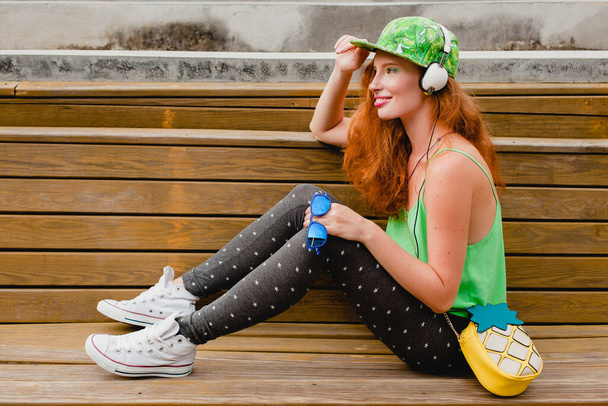 молода стильна жінка з імбиром, що сидить на лавці, слухає музику, навушники, зелену шапку, легінси, сонцезахисні окуляри, весело, модний одяг, гаманець
 - Фото, зображення